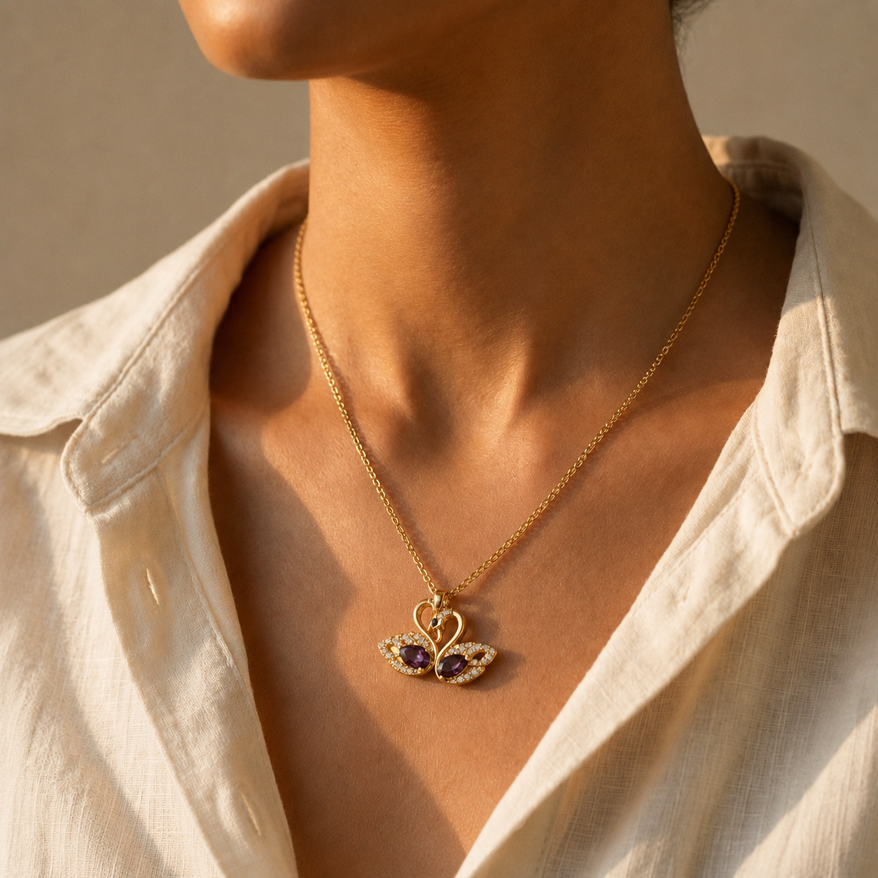 A close-up of a model wearing a gold toned amora teddy demi fine chain necklace with a swan pendant featuring purple gemstone wings, styled with an off-white linen shirt.