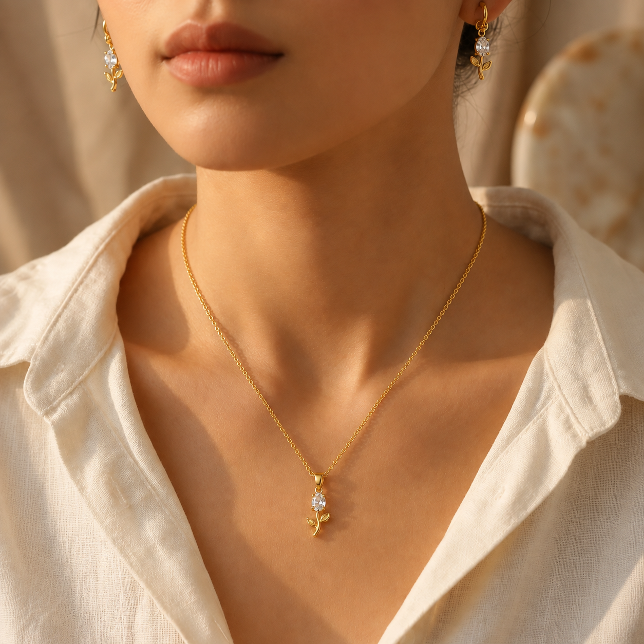 A close-up lifestyle shot of a model wearing a delicate gold plated necklace with a small floral pendant featuring a clear crystal, styled with matching gold earrings and a cream-colored linen shirt.