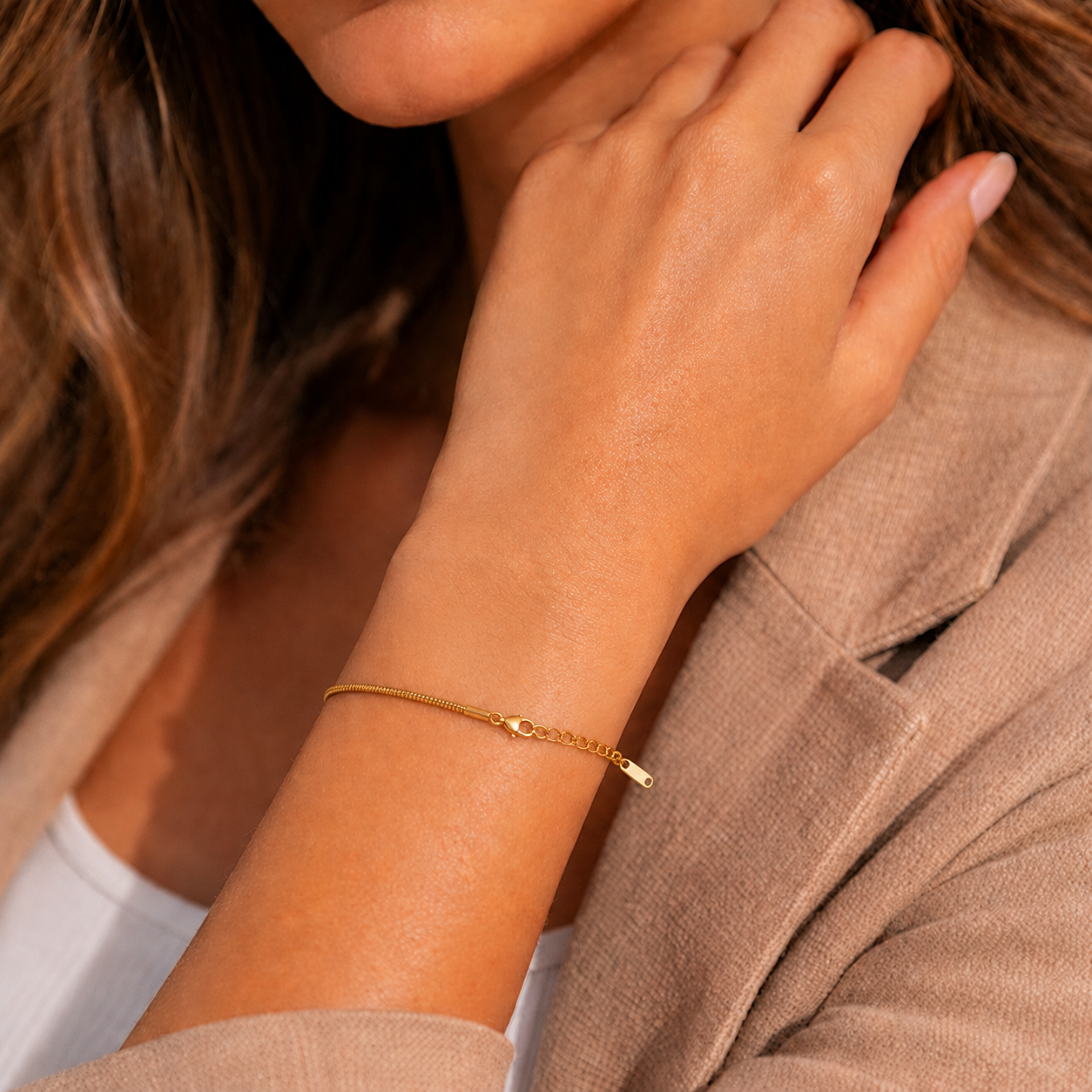 A delicate gold polished chain bracelet with a small polished gold charm, worn on a woman's wrist. The photo is a close-up lifestyle shot showing the jewelry on a model wearing a beige blazer.