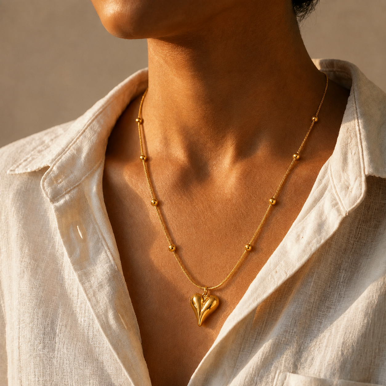 A close-up of a model wearing a delicate gold toned necklace with small bead accents and a heart-shaped pendant, styled with an open white linen shirt.