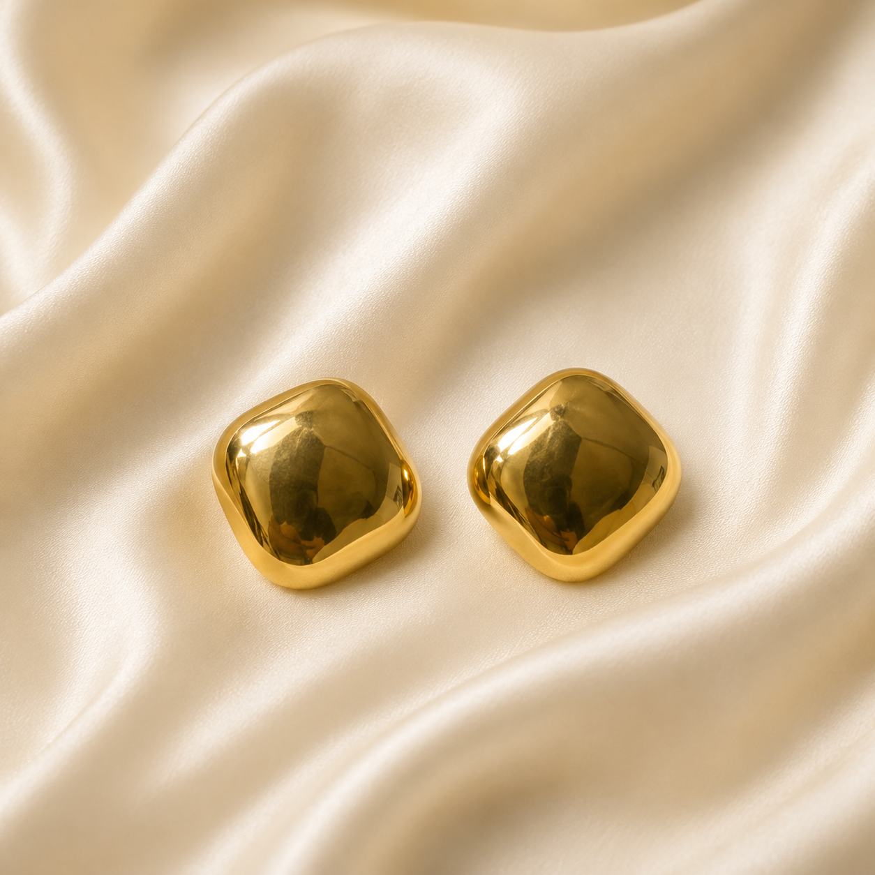 A pair of shiny gold square stud earrings with rounded corners, resting on draped cream satin fabric.