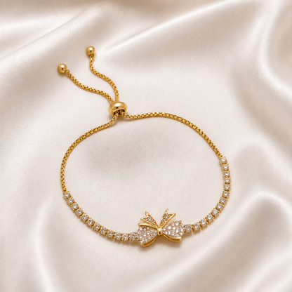 A gold tonned adjustable bracelet with a bow-shaped centerpiece paved with clear stones, displayed on a draped cream satin fabric background.