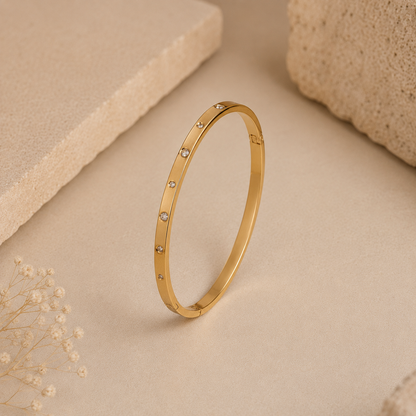 A slim gold tonned bangle bracelet with small clear gemstones spaced along the front, photographed standing upright on a warm beige surface with decorative stone and dried flower props.