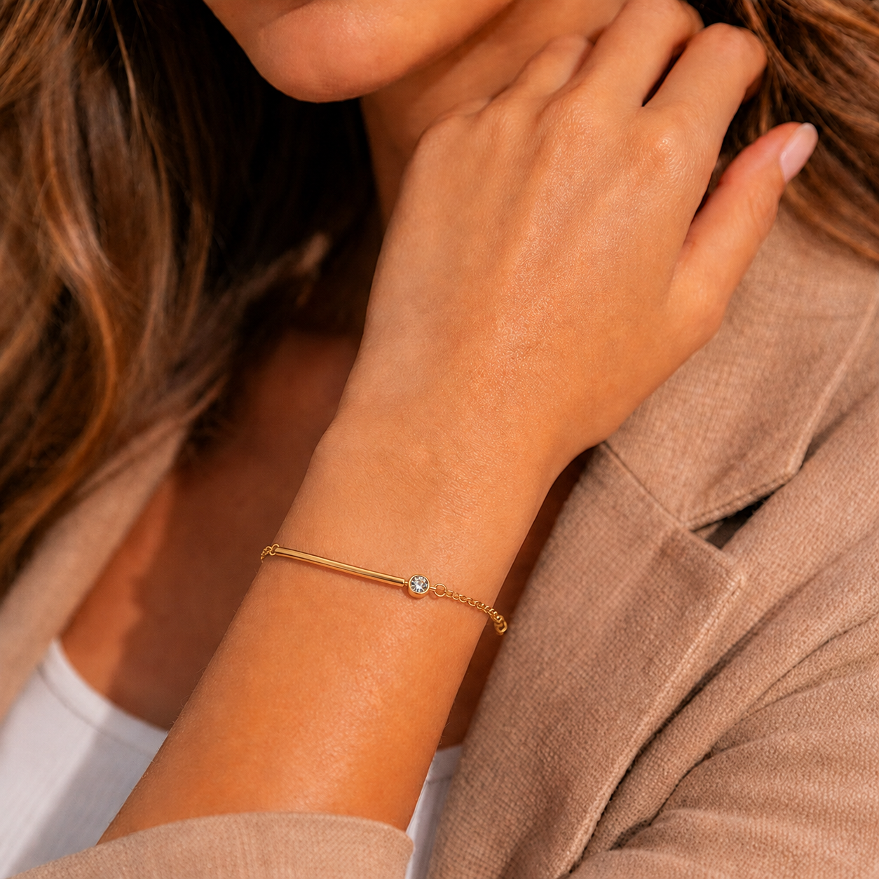 A close-up of a delicate gold tonned chain bracelet with a single small clear gemstone, worn on a woman's wrist. The person is wearing a beige blazer and touching their neck.