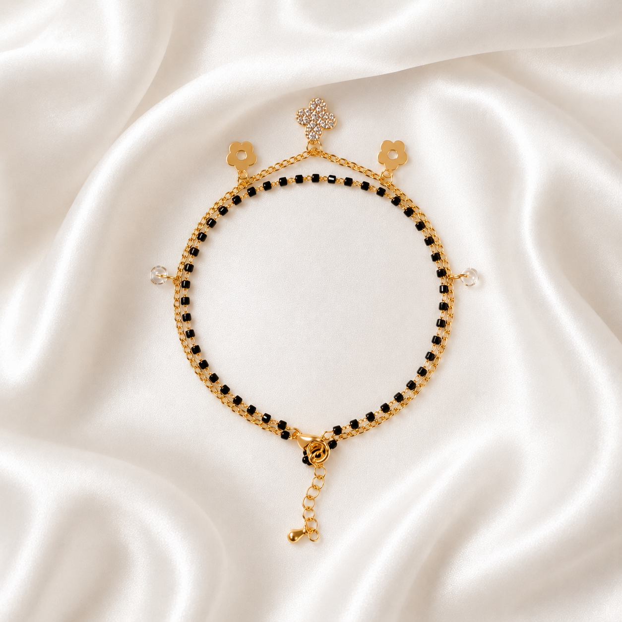 A delicate gold tonned chain bracelet with small black beads, floral charms, and a dangling chain extension, displayed on a draped white satin fabric background.
