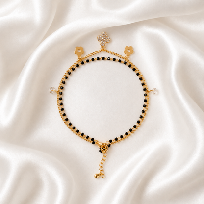 A delicate gold tonned chain bracelet with small black beads, floral charms, and a dangling chain extension, displayed on a draped white satin fabric background.