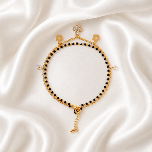 A delicate gold tonned chain bracelet with small black beads, floral charms, and a dangling chain extension, displayed on a draped white satin fabric background.