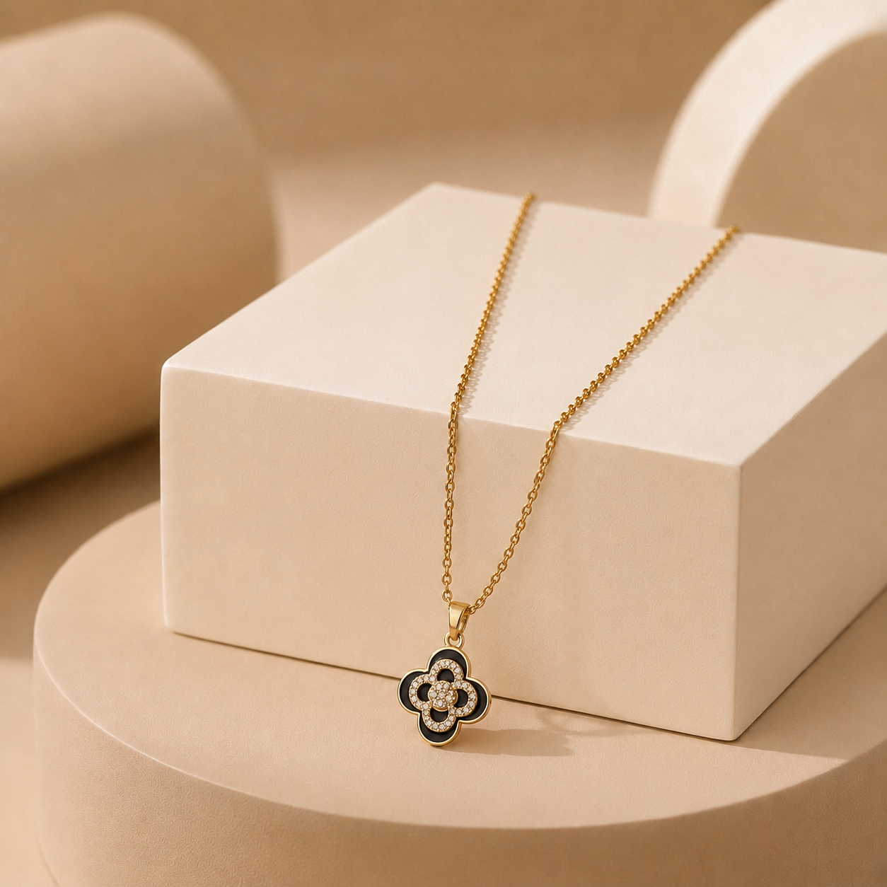 A gold tonned chain necklace with a clover-shaped pendant, featuring black enamel and clear stones, displayed on a cream-colored geometric pedestal.