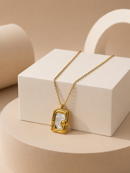 A gold tonned chain necklace with a geometric, open-frame pendant featuring a white inlay, displayed on a white geometric pedestal against a warm beige background.