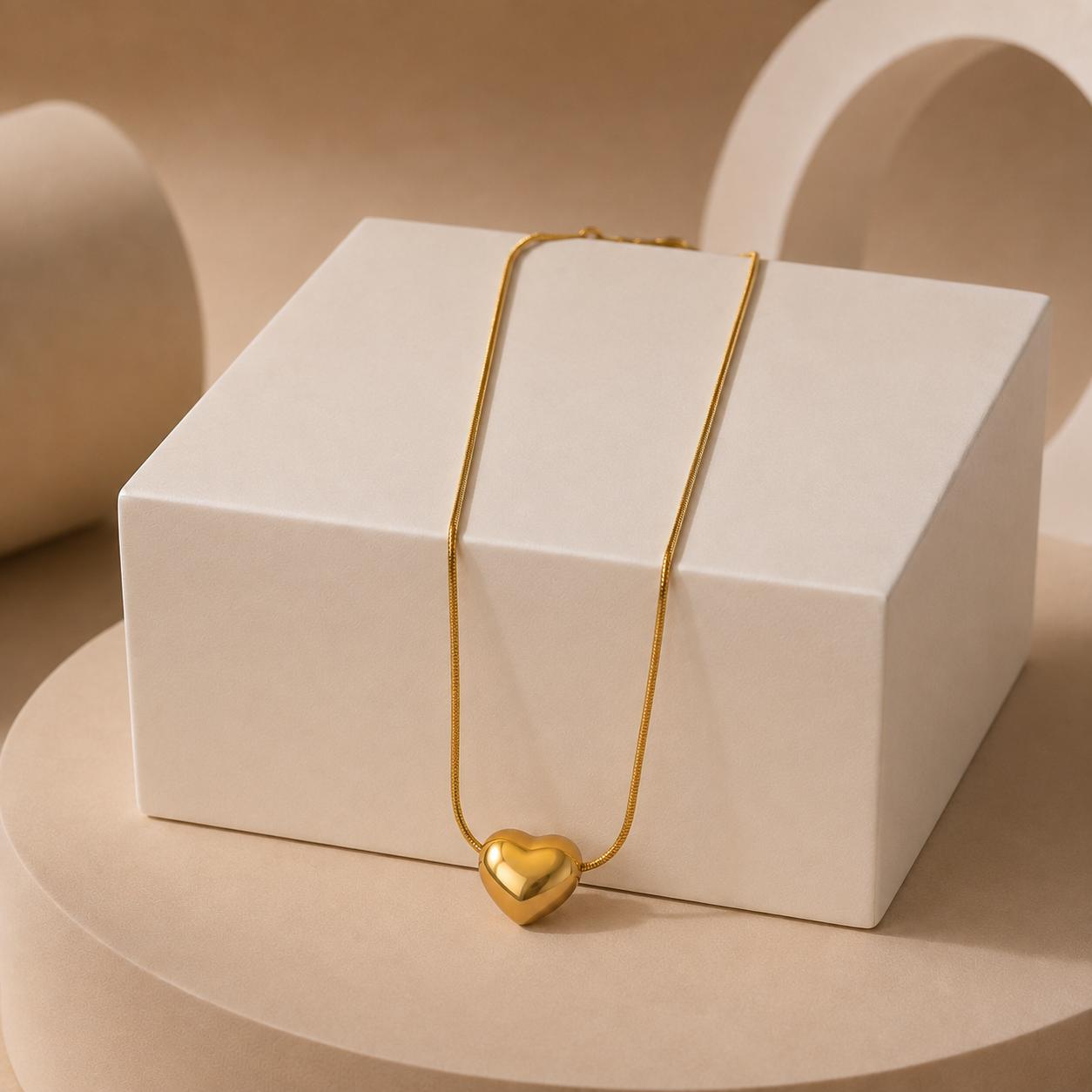 A delicate gold tonned chain necklace with a polished gold heart pendant, draped over a white rectangular display box on a beige pedestal.