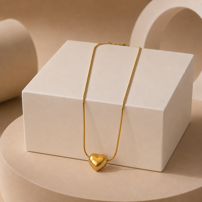 A delicate gold tonned chain necklace with a polished gold heart pendant, draped over a white rectangular display box on a beige pedestal.