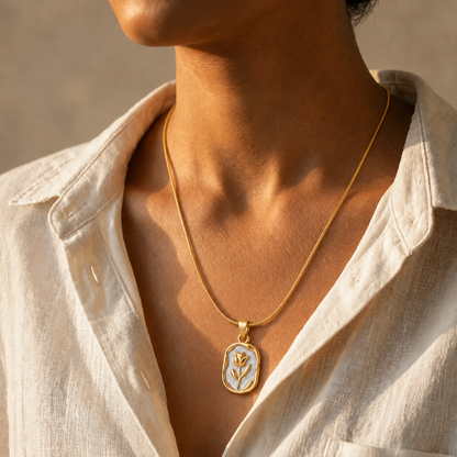 A gold tonned chain necklace with a small oval pendant featuring a white background and a gold flower design, worn by a model in a light-colored linen shirt.