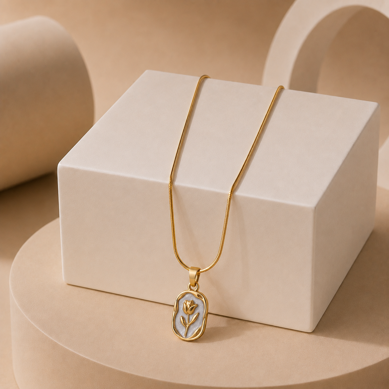 A gold tonned chain necklace with an oval pendant featuring a gold flower design on a light blue background, displayed on a white geometric block against a warm beige studio backdrop.