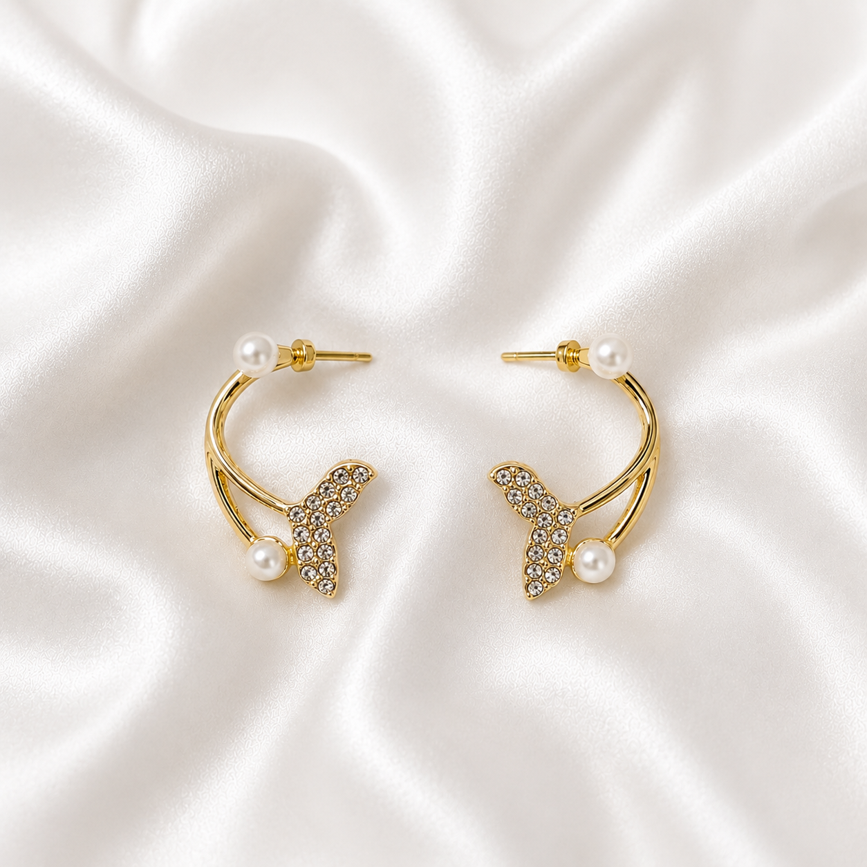 A pair of gold tonned crescent hoop earrings with butterfly-shaped crystal embellishments and small white pearls, displayed on a white satin fabric background.