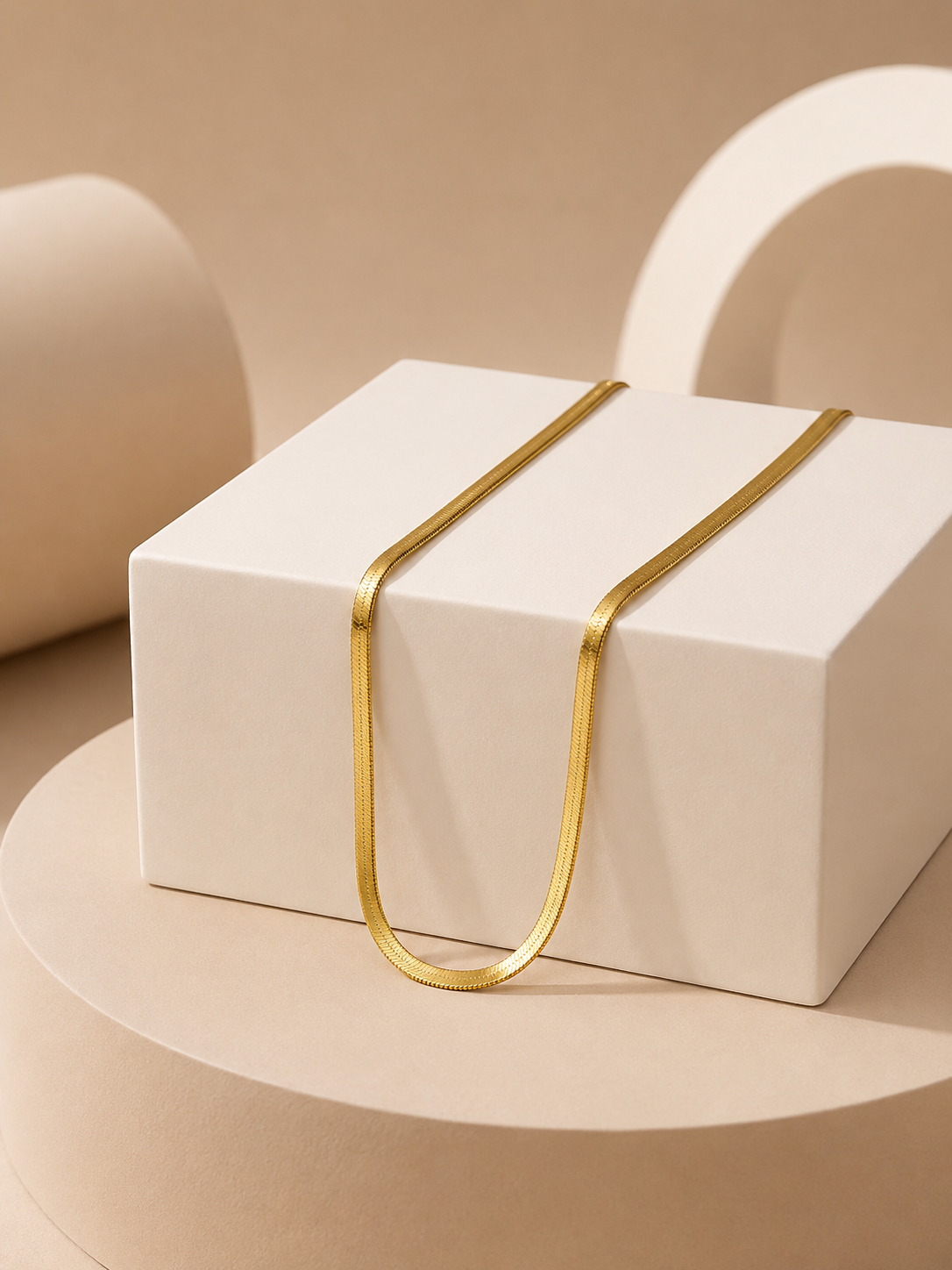 A minimalist gold tonned metal necklace draped over a white geometric display box, set against a warm beige background with abstract 3D shapes.