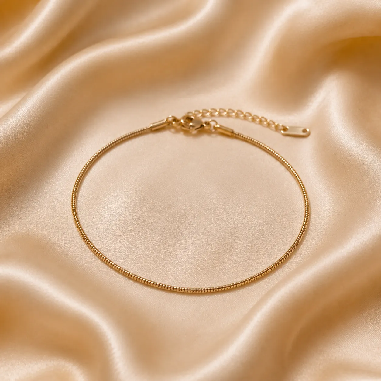 A delicate gold tonned snake chain bracelet with a lobster clasp and extension chain, displayed on a draped beige satin fabric background.