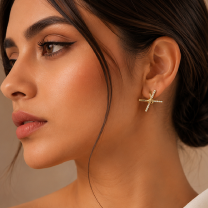 A close-up side profile of a woman wearing a small gold cross stud earring adorned with tiny clear stones, set against a soft beige background.