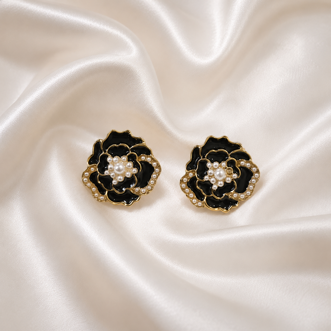 A pair of black and gold flower-shaped earrings with white pearl centers, displayed on a draped cream satin fabric background.