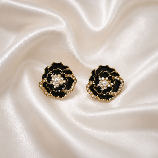 A pair of black and gold flower-shaped earrings with white pearl centers, displayed on a draped cream satin fabric background.