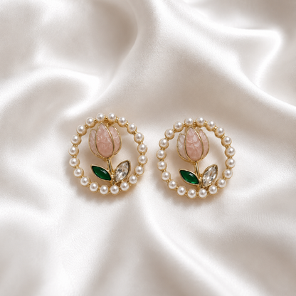 A pair of gold-tone floral stud earrings with a border of white faux pearls, featuring a pink stone flower, green stone leaves, and clear marquise crystals, displayed on a draped white satin fabric.