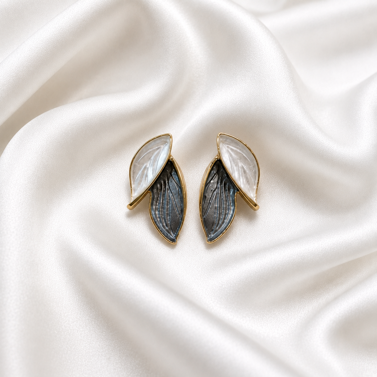 A pair of gold-tone leaf-shaped earrings with one white mother-of-pearl leaf and one textured gray-blue leaf, displayed on a draped white satin fabric background.
