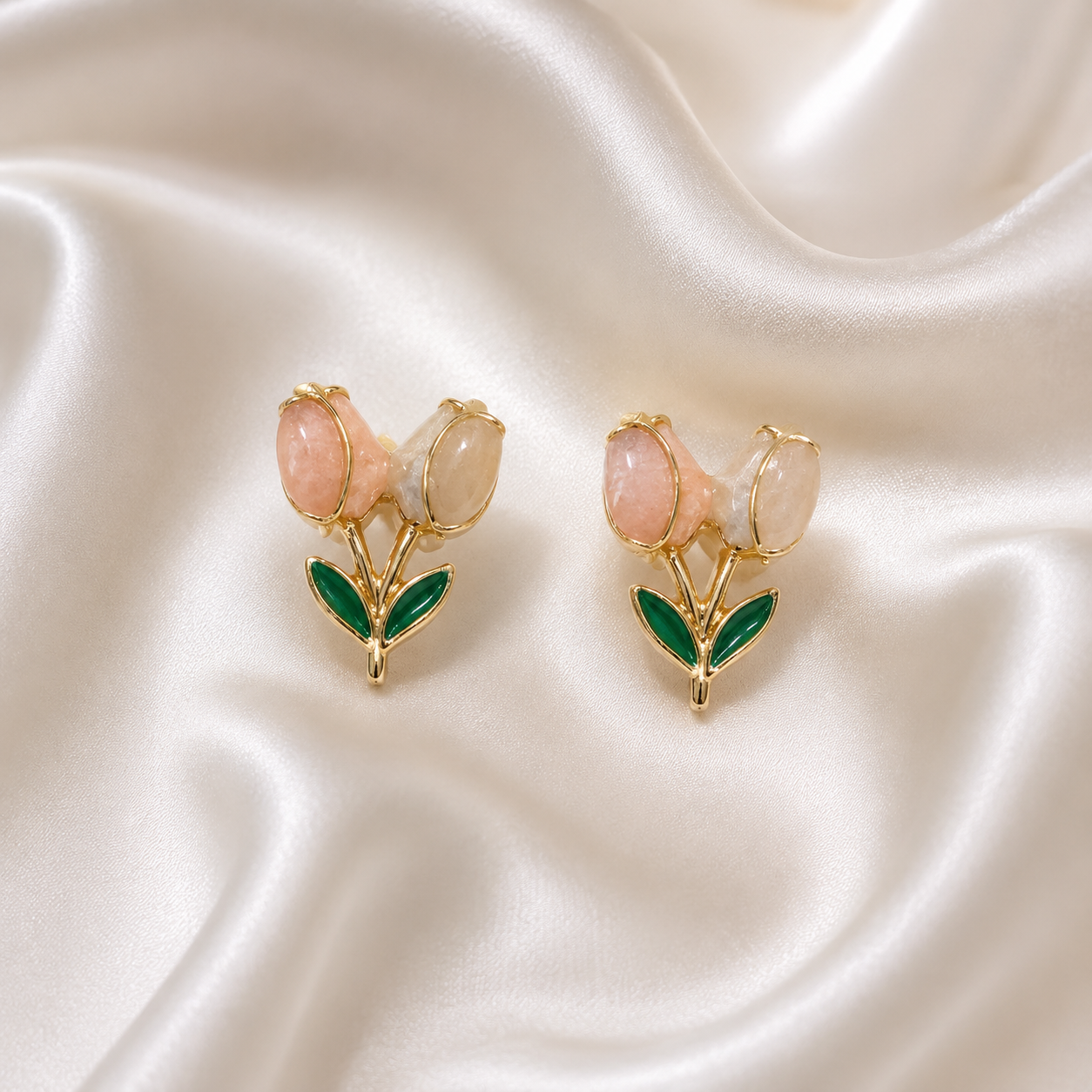 A pair of gold-tone stud earrings designed as tulips, featuring pink and milky white stones with green enamel leaves, displayed on a draped white satin fabric.