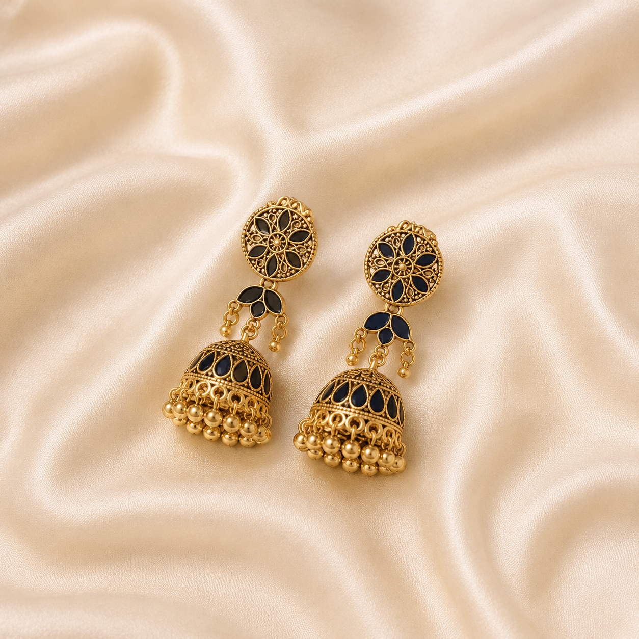 A pair of gold-toned Indian jhumka earrings with black enamel detailing, displayed on a draped beige satin fabric background.