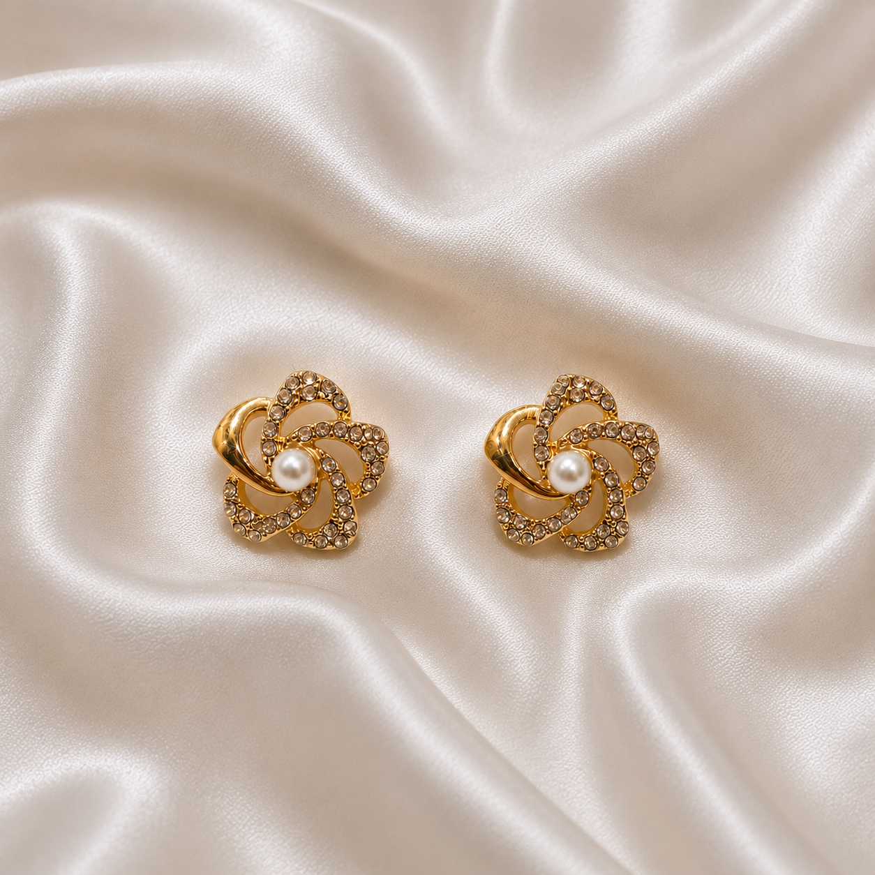 A pair of gold-toned flower-shaped earrings with a central white pearl and small clear stones, displayed on a draped cream satin fabric background.