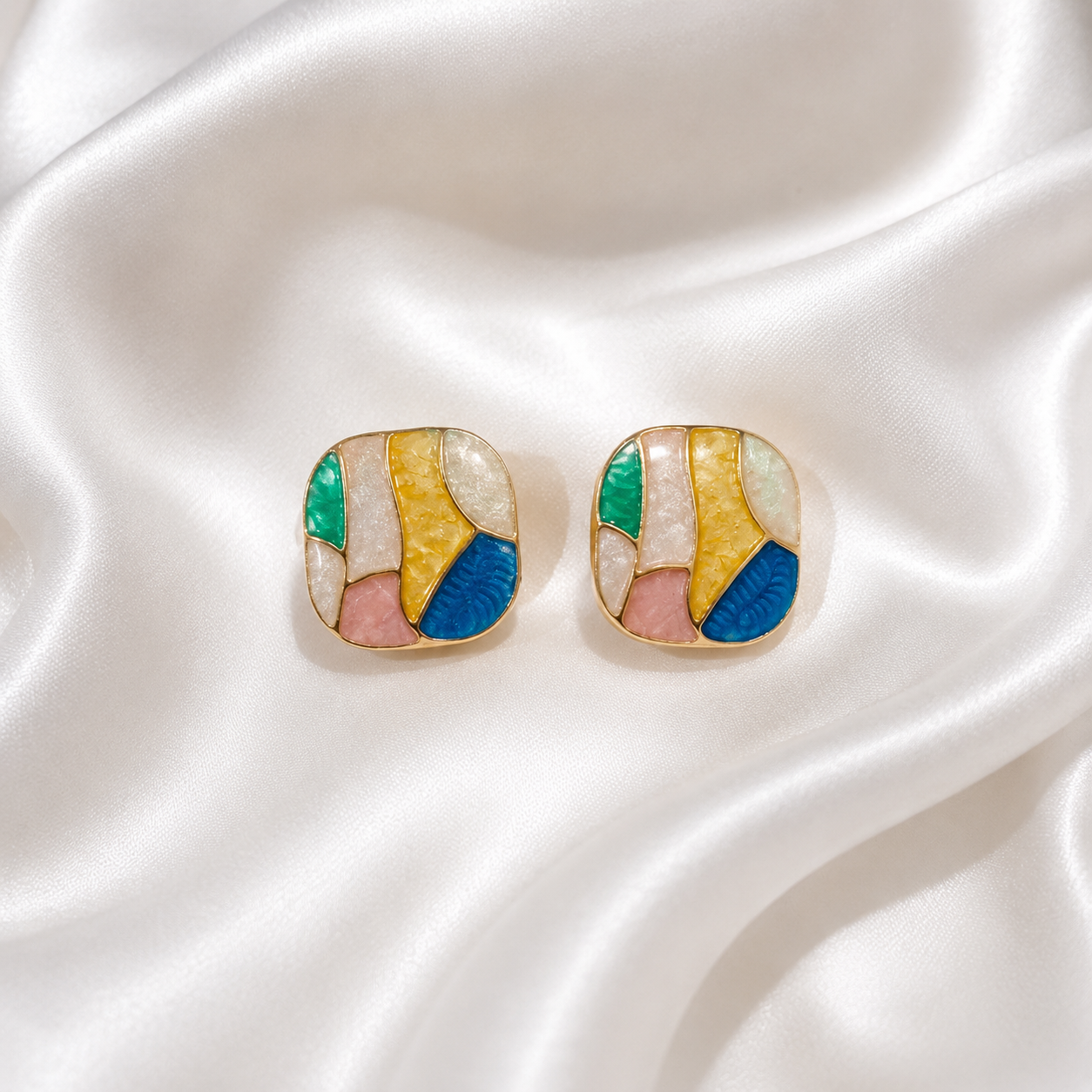 A pair of gold-toned square stud earrings with an abstract mosaic design in yellow, blue, pink, green, and white, displayed on a draped white satin fabric.