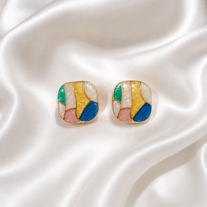 A pair of gold-toned square stud earrings with an abstract mosaic design in yellow, blue, pink, green, and white, displayed on a draped white satin fabric.