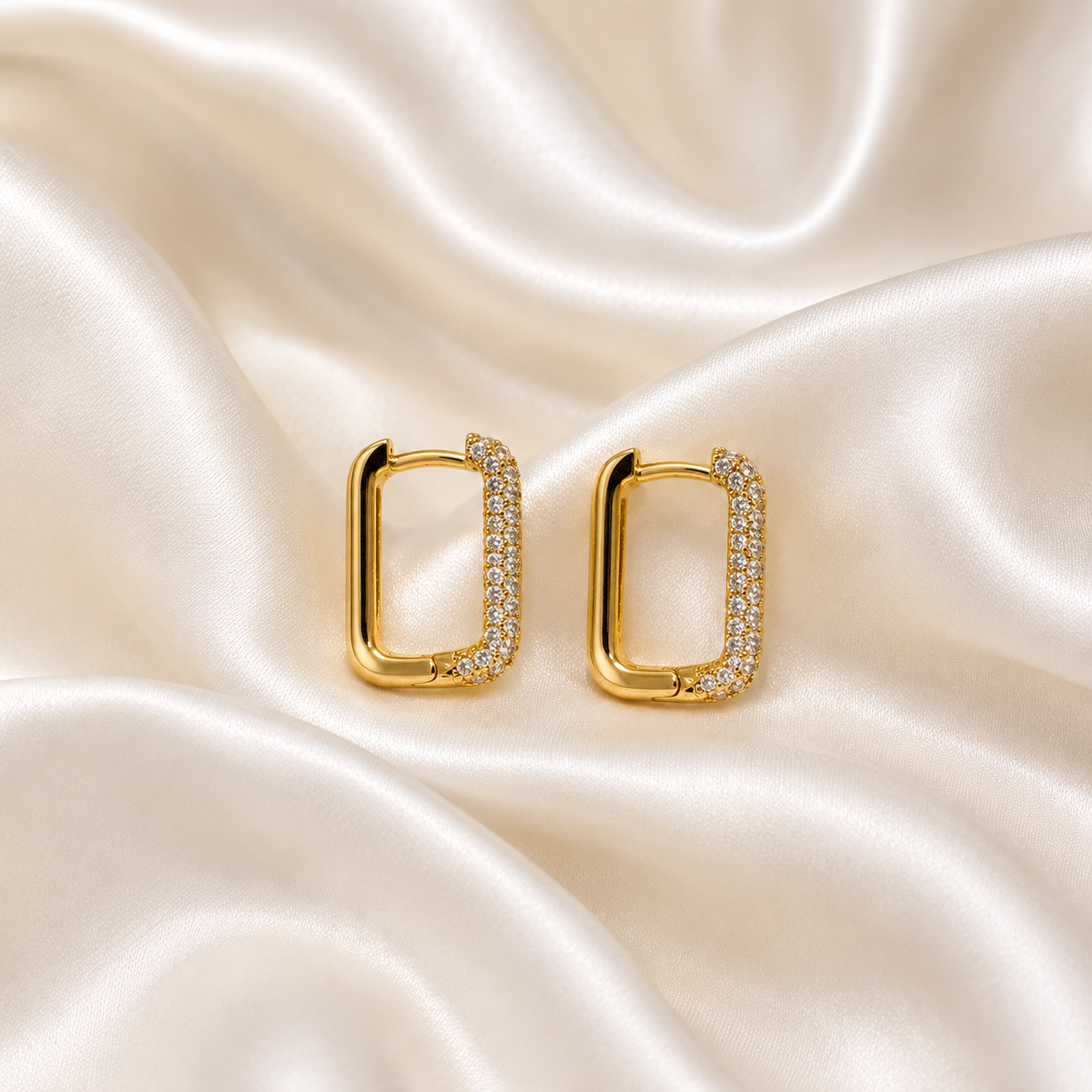 A pair of gold rectangular hoop earrings with pavé-set clear stones, displayed on a draped cream satin fabric background.