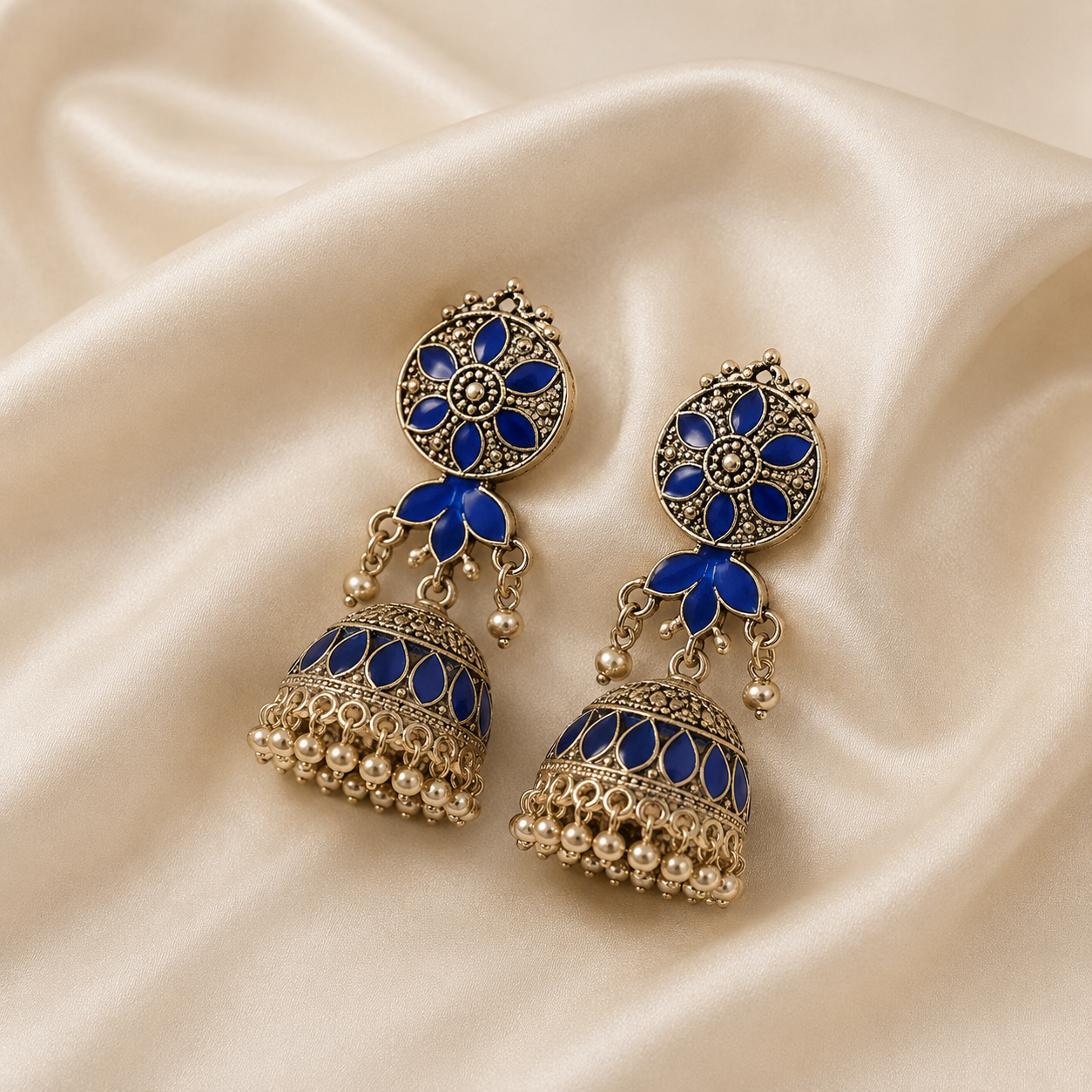 A pair of ornate Indian jhumka earrings with blue enamel and gold-toned filigree, displayed on a draped cream satin fabric background.