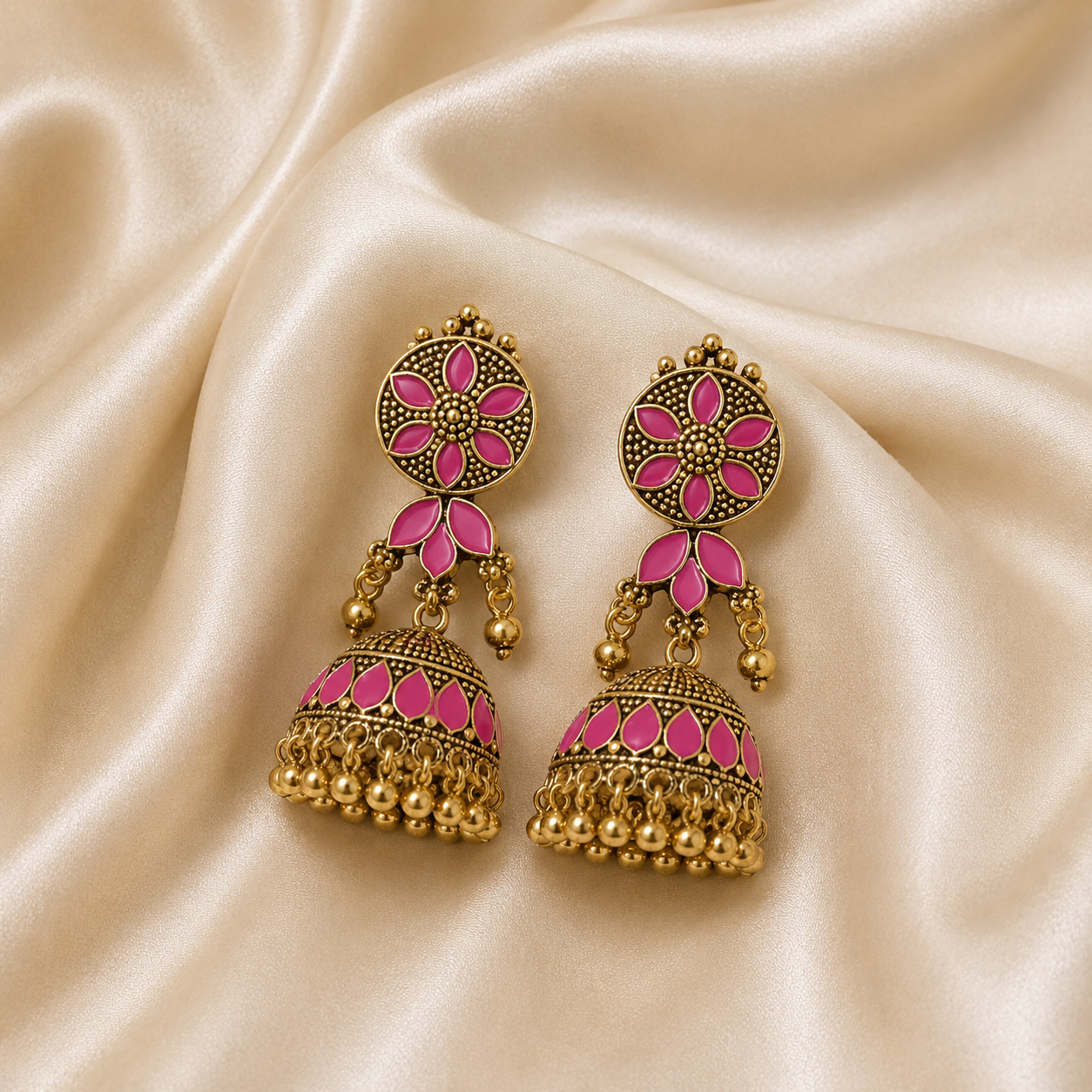 A pair of ornate gold-toned jhumka earrings with bright pink enamel detailing, displayed on a draped cream satin fabric background.