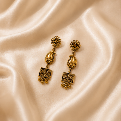 A pair of ornate gold-toned traditional drop earrings with intricate filigree and paisley details, displayed on a draped cream satin fabric background.