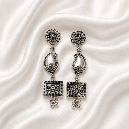 A pair of ornate silver-toned dangle earrings with intricate filigree detailing, displayed on a draped white satin fabric background.