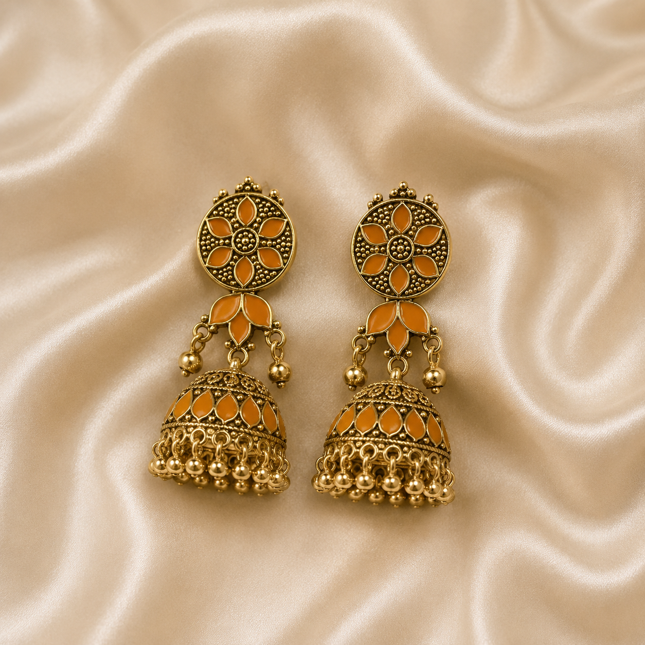 A pair of traditional Indian gold-plated jhumka earrings with orange enamel detailing, displayed on a draped beige satin fabric background.