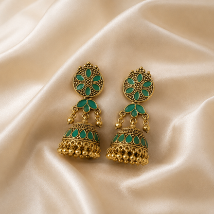 A pair of traditional Indian gold-plated jhumka earrings with green enamel detailing, displayed on a draped cream satin fabric background.
