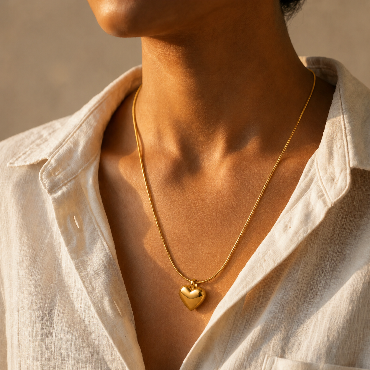 A close-up lifestyle shot of a person wearing a delicate gold chain necklace with a small, polished gold heart pendant. The person is wearing a light beige linen button-up shirt, and the background is a warm, neutral-toned wall.