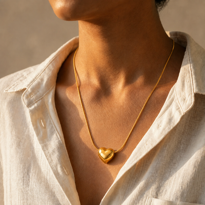 A close-up of a person wearing a delicate gold tonned chain necklace with a polished heart-shaped pendant, styled with a light beige linen shirt under warm sunlight.