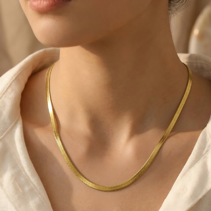 A close-up of a person's neck and upper chest wearing a thin, polished gold tonned snake chain necklace over a white top.