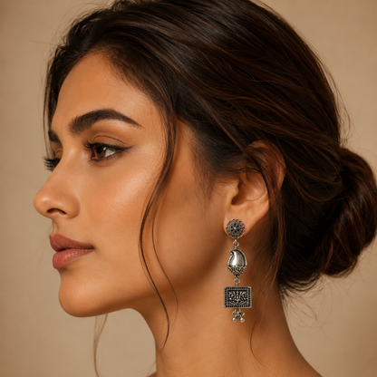 A close-up side profile of a woman wearing a silver-toned traditional Indian jhumka earring with intricate filigree detailing and a dangling square charm.