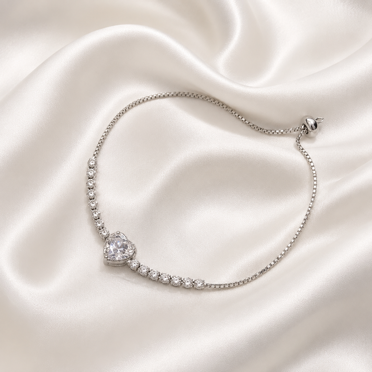 A delicate silver plated chain bracelet with a heart-shaped crystal centerpiece and small clear stones along the curve, displayed on a draped cream satin fabric background.
