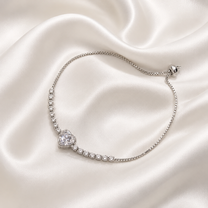A delicate silver plated chain bracelet with a heart-shaped crystal centerpiece and small clear stones along the curve, displayed on a draped cream satin fabric background.