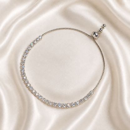 A delicate silver tennis-style necklace with clear sparkling stones, displayed on a draped cream satin fabric background.