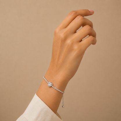 A delicate silver tonned bracelet with a heart-shaped crystal charm and a small dangling bead, shown worn on a woman's wrist against a warm beige background.