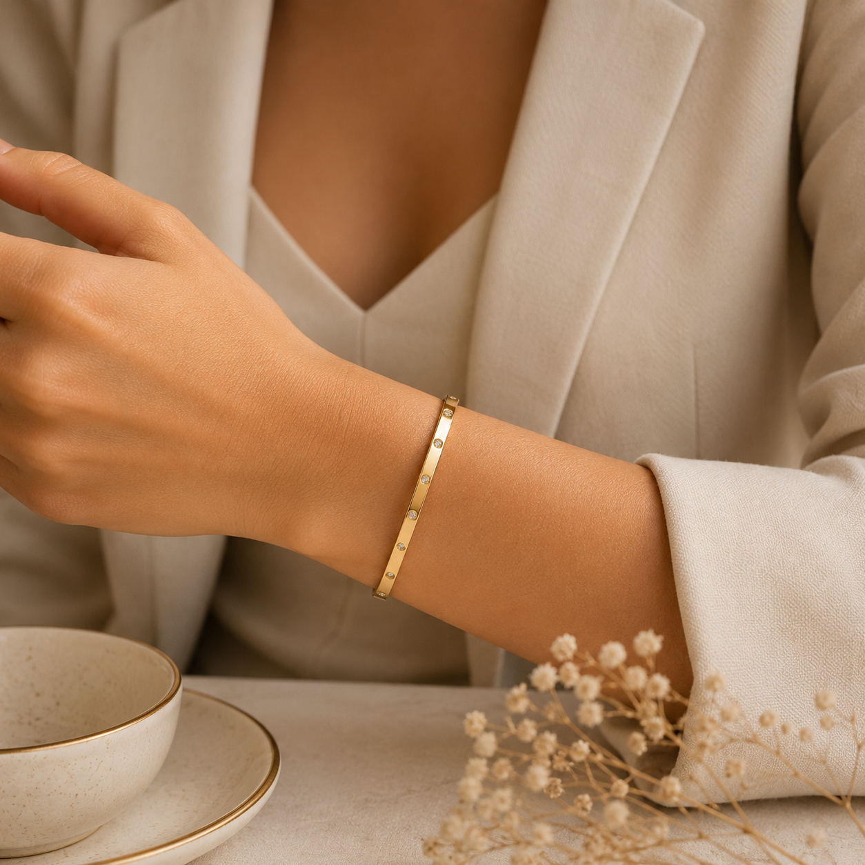 A close-up of a person's wrist wearing a slim gold tonned bangle bracelet, styled with a beige blazer, a teacup, and dried flowers on a table.