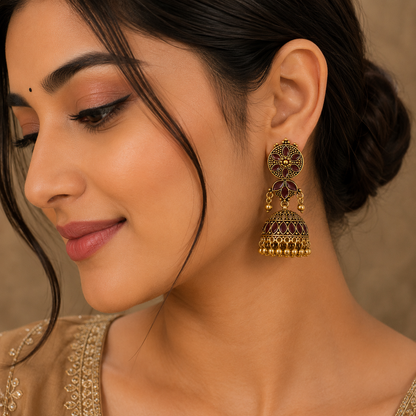 A close-up lifestyle shot of a woman wearing a traditional Indian gold tonned and maroon jhumka earring. The earring features intricate filigree detailing and dangling bell-shaped elements, shown on a model with dark hair tied back.