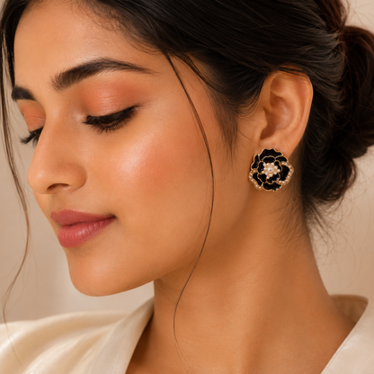 A close-up of a woman wearing a black and gold flower-shaped stud earring with a small clear center stone, shown against a soft beige background.