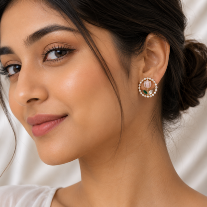 A close-up of a woman wearing a circular gold-toned earring adorned with a pink lotus flower, green leaves, and a border of white pearls.