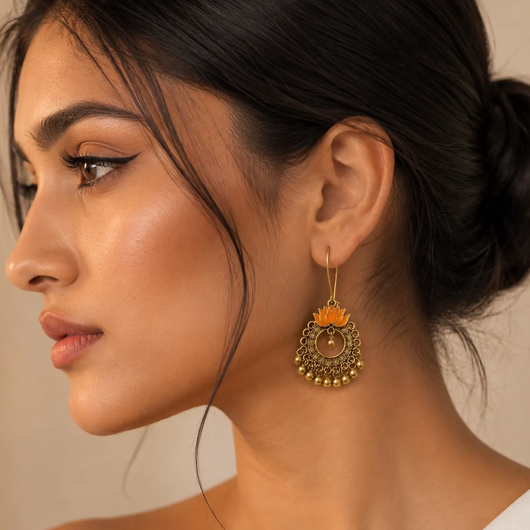A close-up side profile of a woman wearing a dangling gold-toned earring with an orange lotus flower design and small gold beads at the bottom.